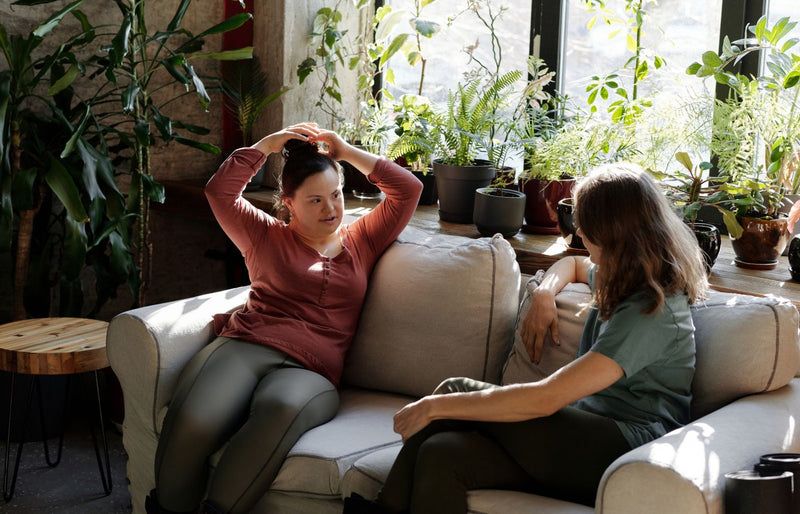 Two women enjoying a relaxed conversation on a sofa in a bright room filled with plants.