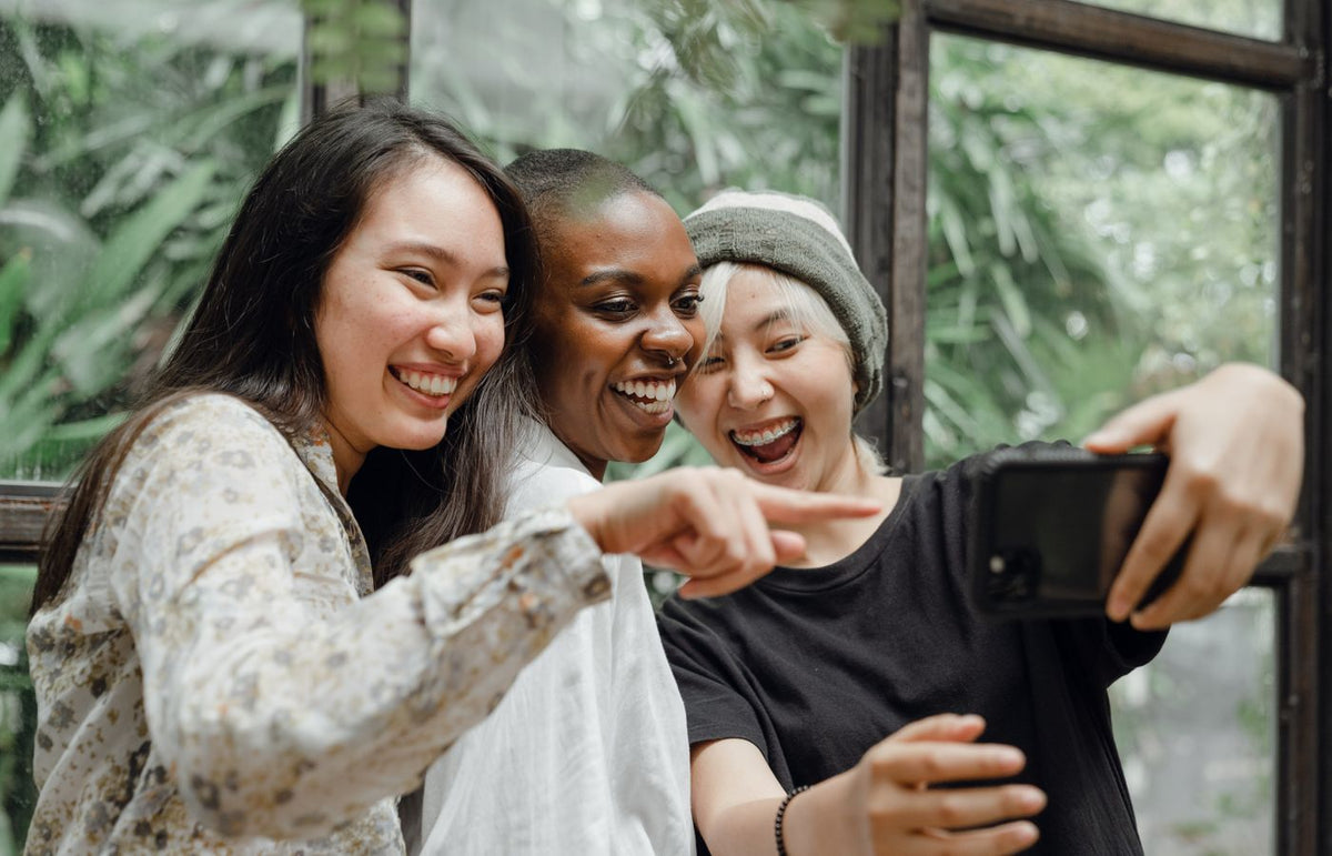 Three friends of different ethnicities laughing and taking a selfie together indoors.