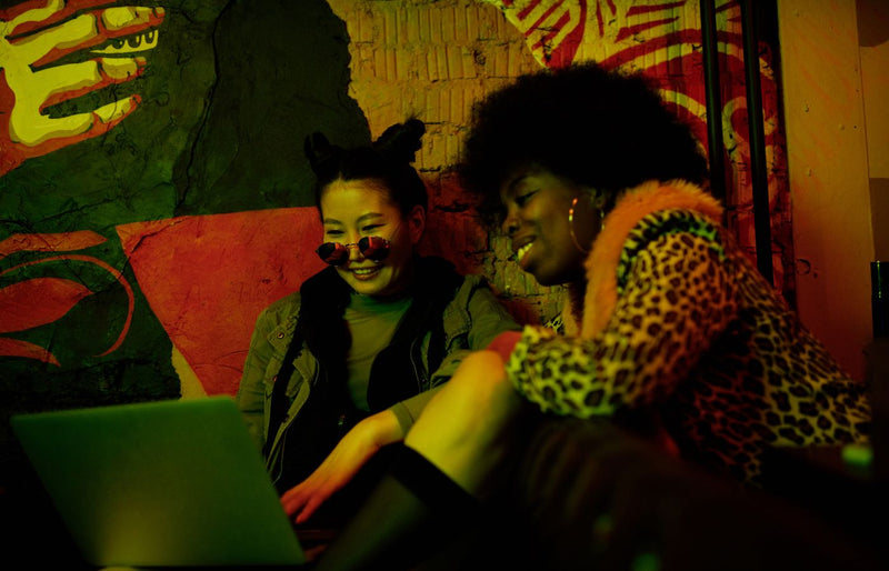 Two fashionable women laughing together while using a laptop in an artistic indoor setting.