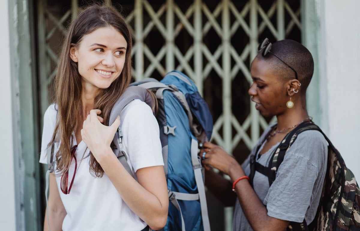 Two women preparing for travel with backpacks, smiling outdoors near a gate.