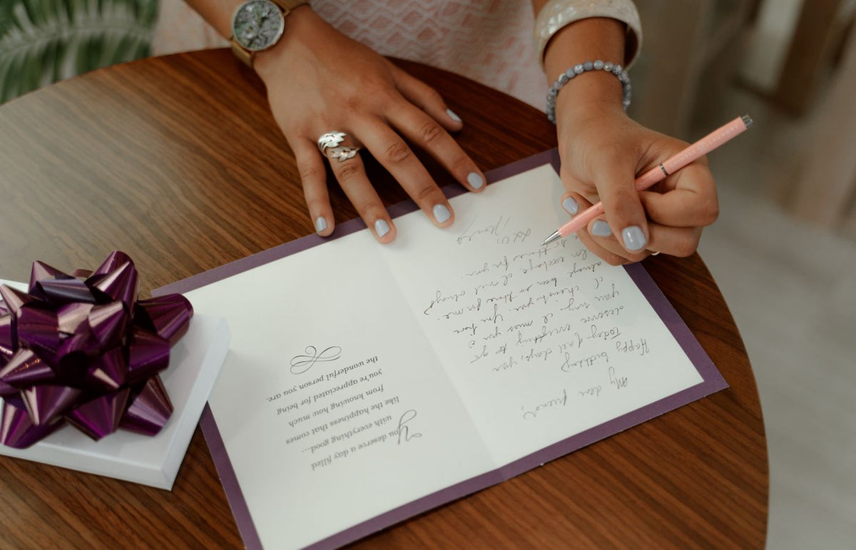 A woman writes a message inside a greeting card on a wooden table with a gift beside.