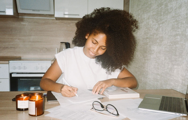 A cheerful woman writing at a home kitchen table with candles, eyeglasses, and a laptop.