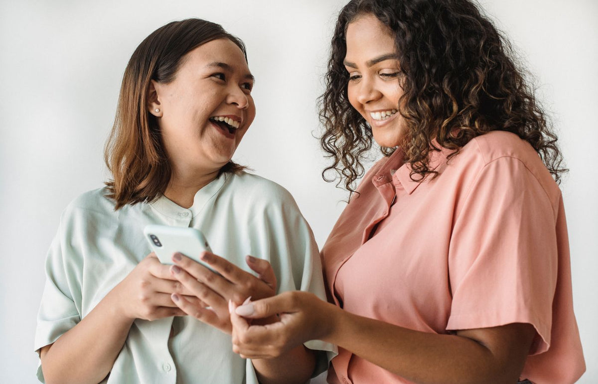 Two women happily sharing a moment while using a smartphone, laughing in a bright indoor setting.