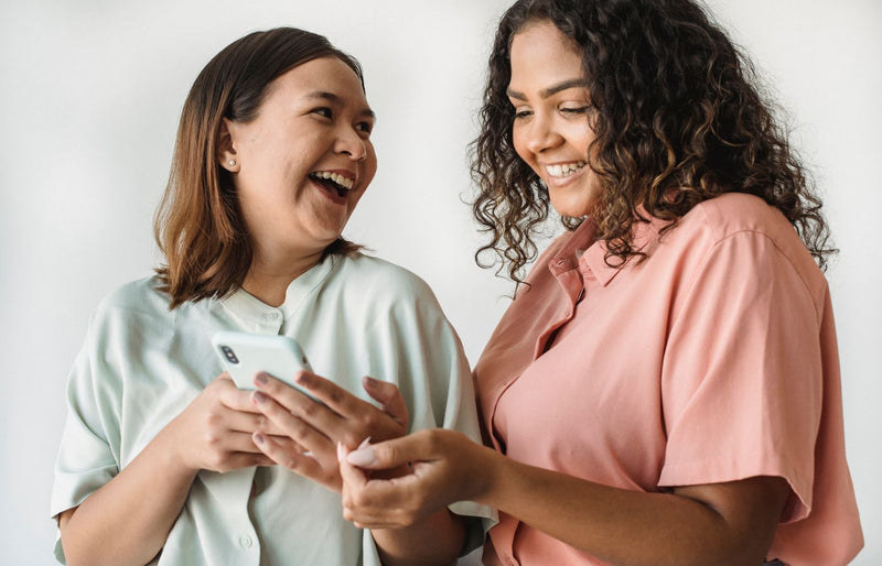 Two women happily sharing a moment while using a smartphone, laughing in a bright indoor setting.