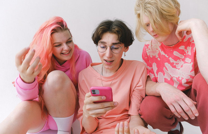 Three happy teenagers sitting together indoors, smiling and looking at a smartphone, enjoying social media or online con