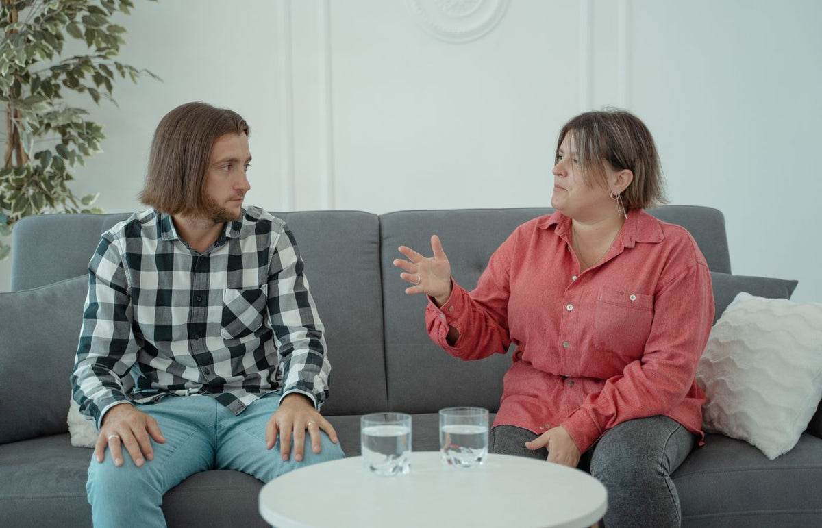 A couple sits on a couch discussing with expressive gestures in a modern indoor setting.
