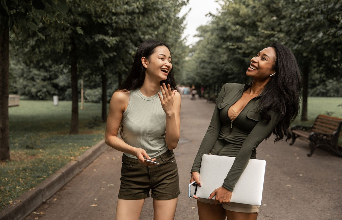 Two young women laugh and enjoy a conversation outdoors in a park on a warm day.