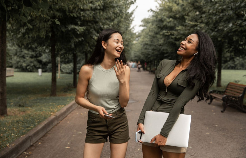 Two young women laugh and enjoy a conversation outdoors in a park on a warm day.