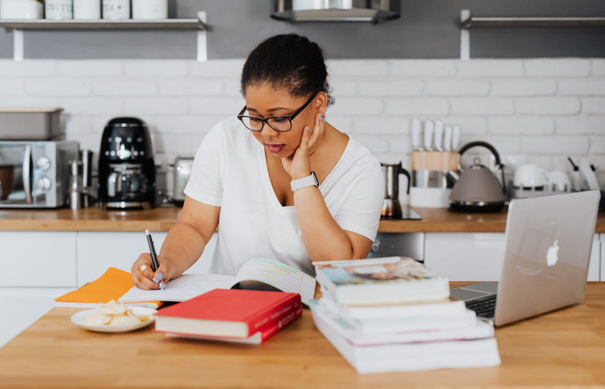 Woman studying at kitchen table surrounded by books and laptop, focused on writing.