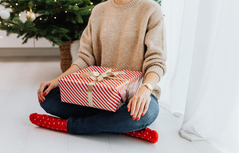 Adult woman holding a wrapped Christmas gift while sitting by a decorated tree indoors.