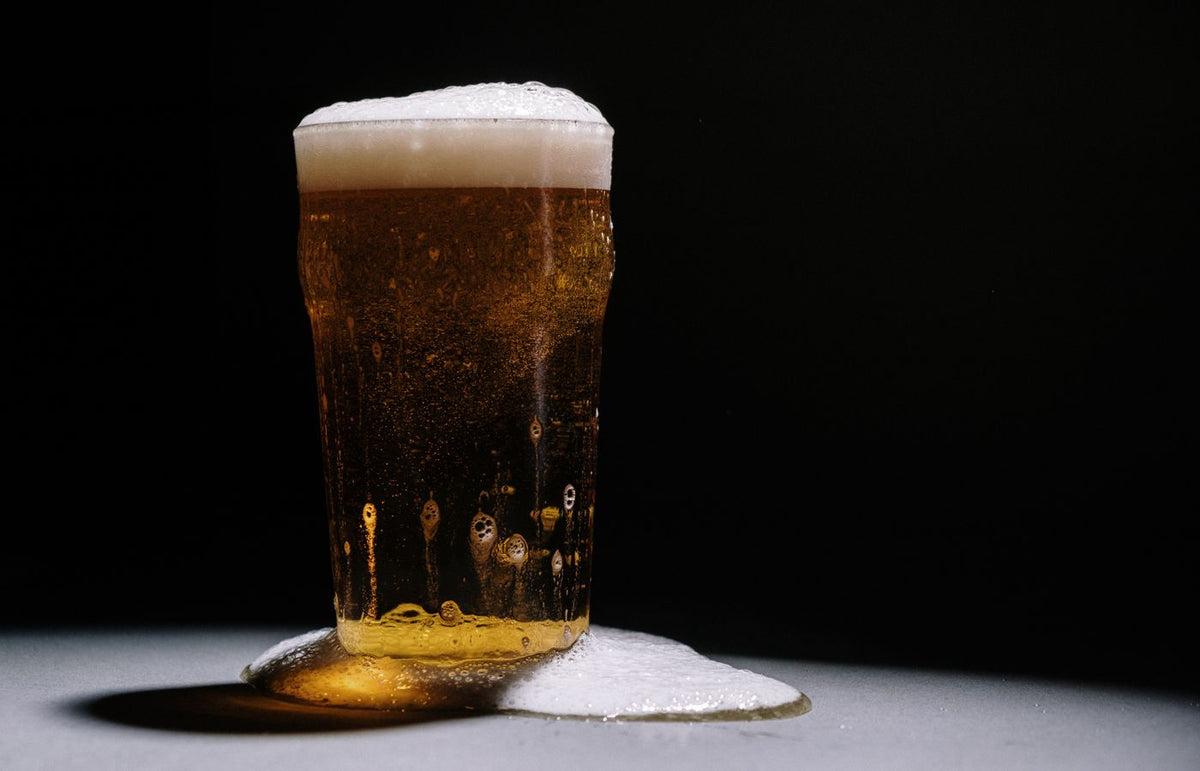 A studio shot of a frothy and overflowing pint of golden beer against a dark backdrop.