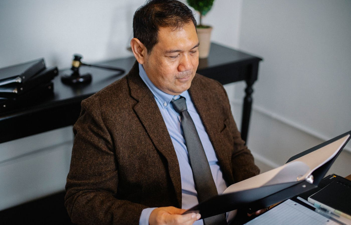 A confident Asian man in formal attire examines documents at his office desk.
