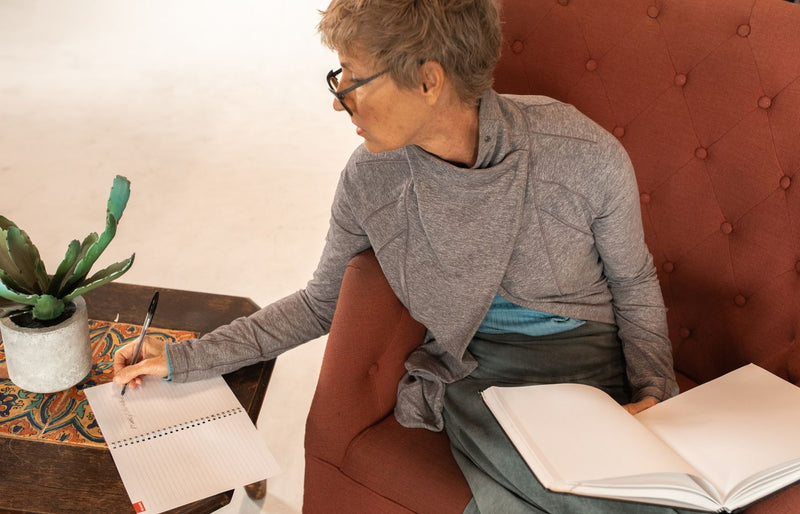 Elderly woman writing notes with a notebook while seated comfortably indoors.