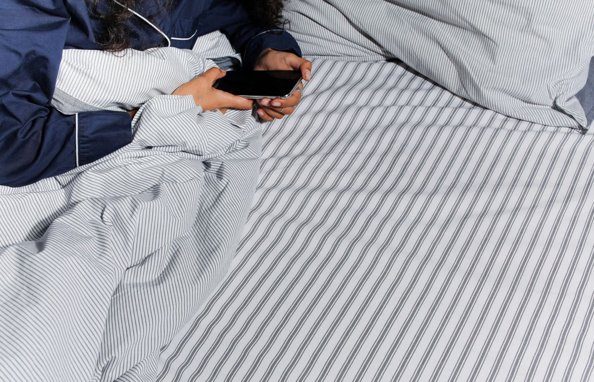 Adult in pajamas using a smartphone in bed with striped bedsheets.