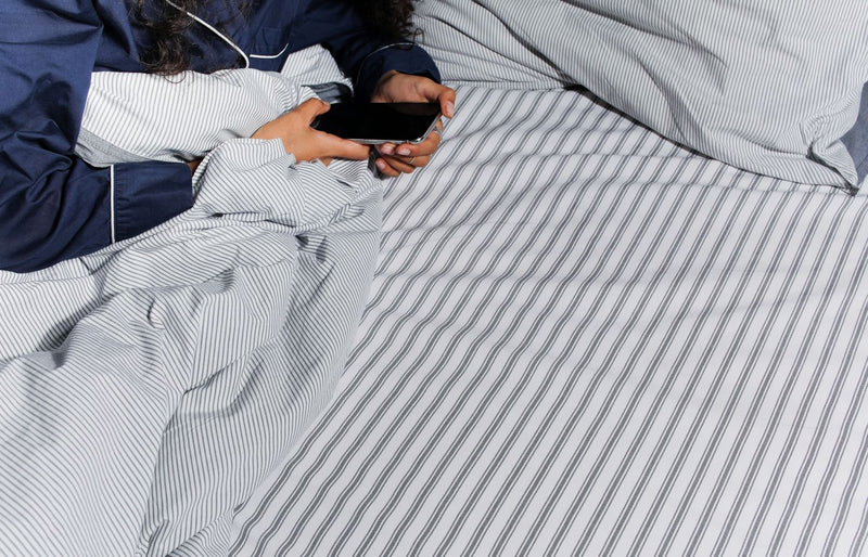 Adult in pajamas using a smartphone in bed with striped bedsheets.