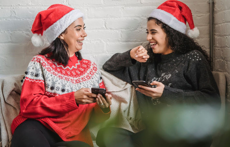 Two women wearing Santa hats enjoying Christmas on their phones, sharing the festive spirit.