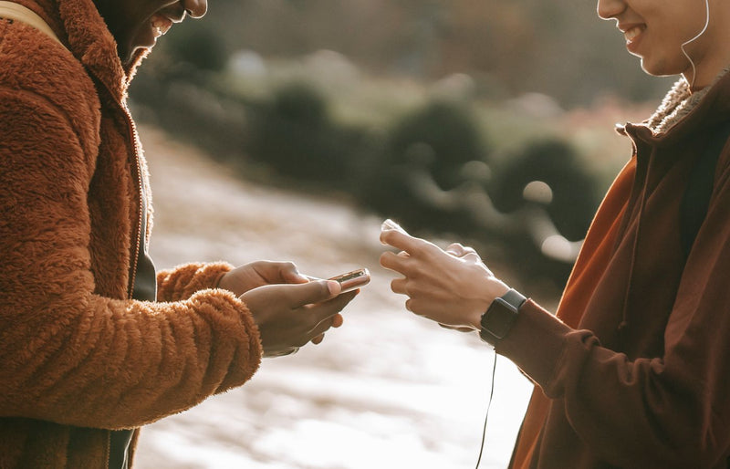 Two friends interacting outdoors, using smartphones in a cheerful, relaxed setting.
