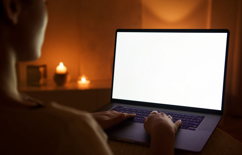 A person working on a laptop with a blank screen, illuminated by candlelight.