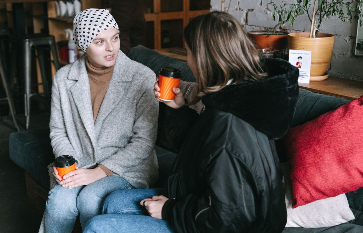 Two women in winter clothing chatting over coffee on a couch indoors.