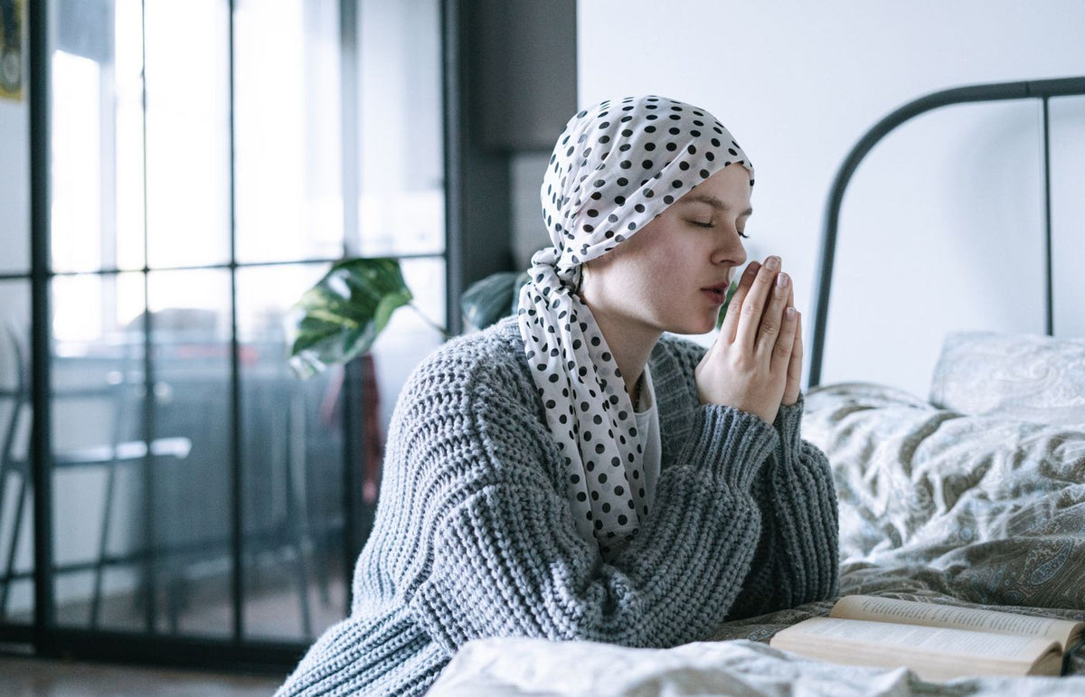 A hopeful woman in a scarf praying in a cozy bedroom during her cancer recovery journey.