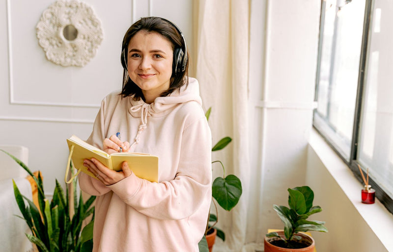 Young woman in a hoodie with headphones writing in a notebook indoors.