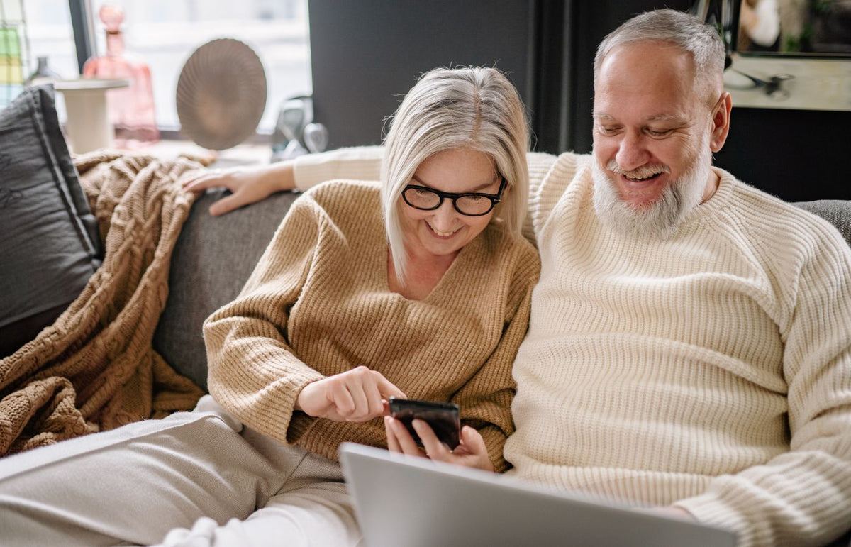 Smiling elderly couple sitting on sofa, engaging with smartphone. Cozy indoor setting.