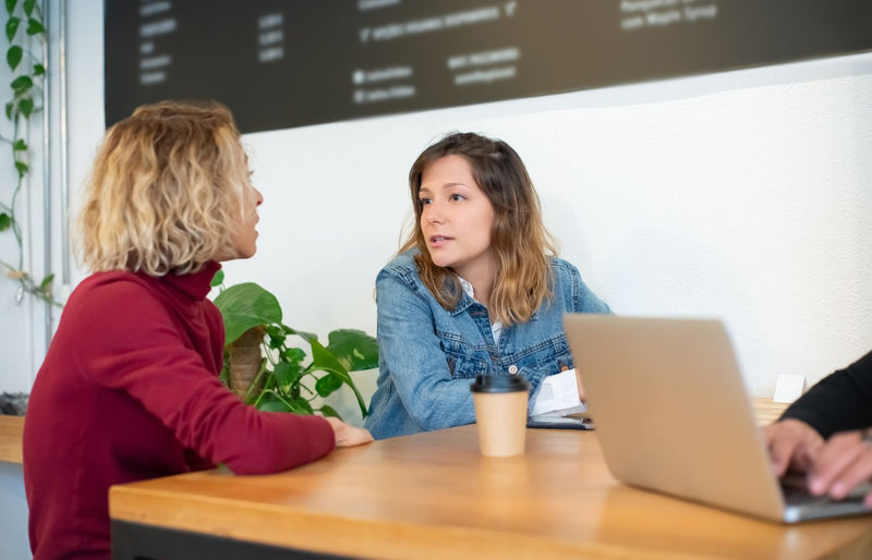 Two women engaging in a casual discussion at a café, seated at a wooden table with drinks and a laptop.