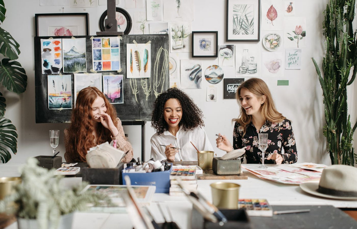 Three women enjoying a painting workshop together surrounded by art supplies and watercolor artwork.
