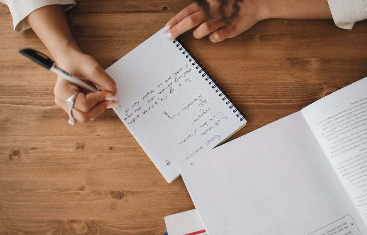 Close-up view of a person taking notes in a notebook with an open book on a wooden desk.