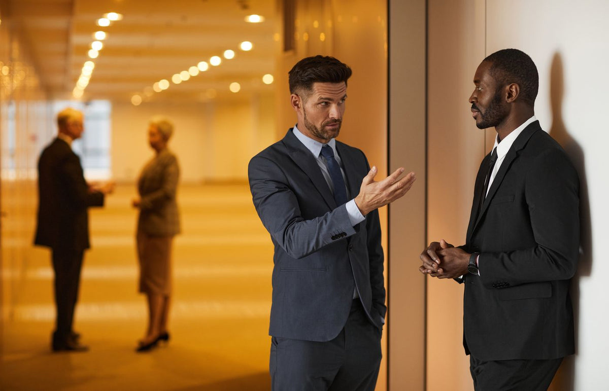 Two businessmen in formal attire engaged in a serious discussion in a modern office hallway.
