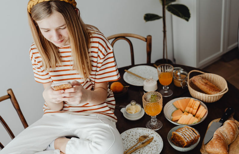 Woman sitting at a dining table using phone, enjoying a home-cooked breakfast with fresh fruits and juice.
