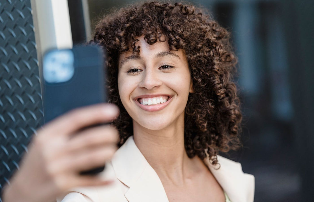 Happy woman with curly hair taking a selfie with a smartphone outdoors, showcasing joy and charisma.