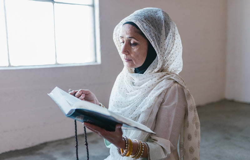 A woman in a hijab reading a Quran and holding prayer beads in a serene indoor setting.