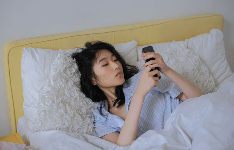 Asian woman comfortably lying in bed, using her phone in a serene bedroom setting.