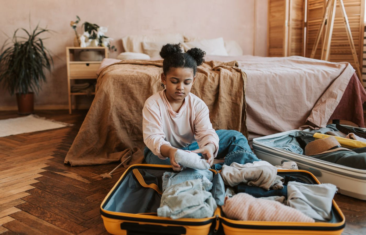 A young girl folding clothes into a suitcase in a cozy bedroom setting with natural decor.