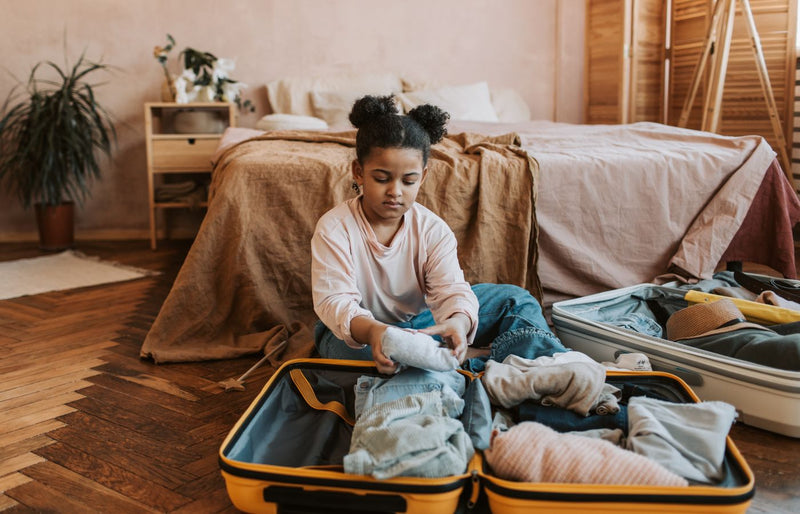A young girl folding clothes into a suitcase in a cozy bedroom setting with natural decor.