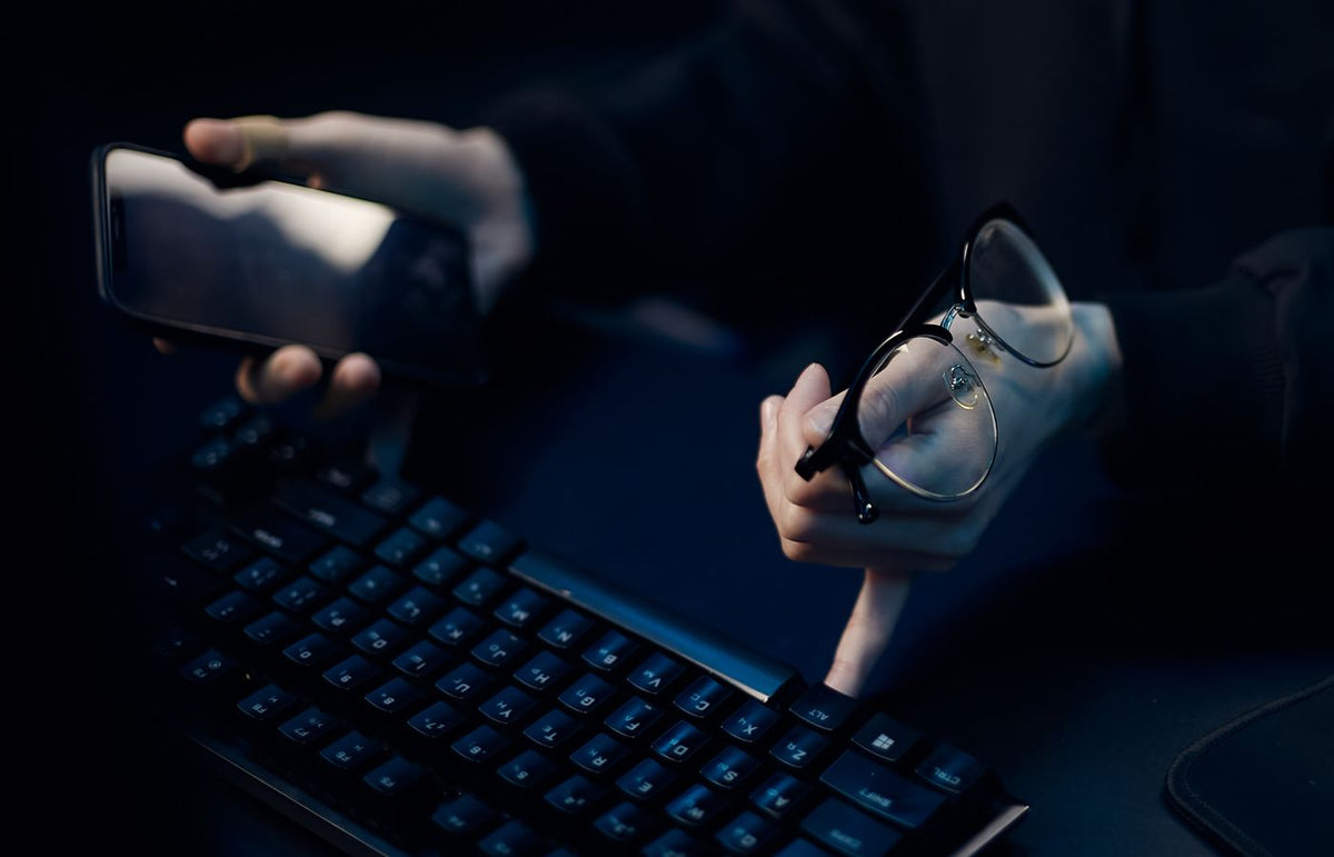 Close-up of hands using smartphone and computer keyboard, holding eyeglasses, focused workspace.