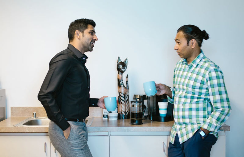 Two colleagues enjoy a coffee break in a modern office kitchen setting.