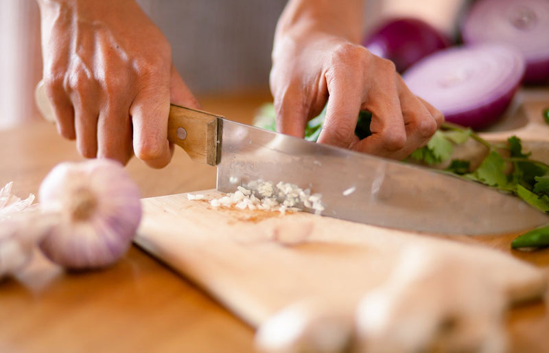 Hands chopping fresh garlic and onions on a wooden board in a kitchen setting.