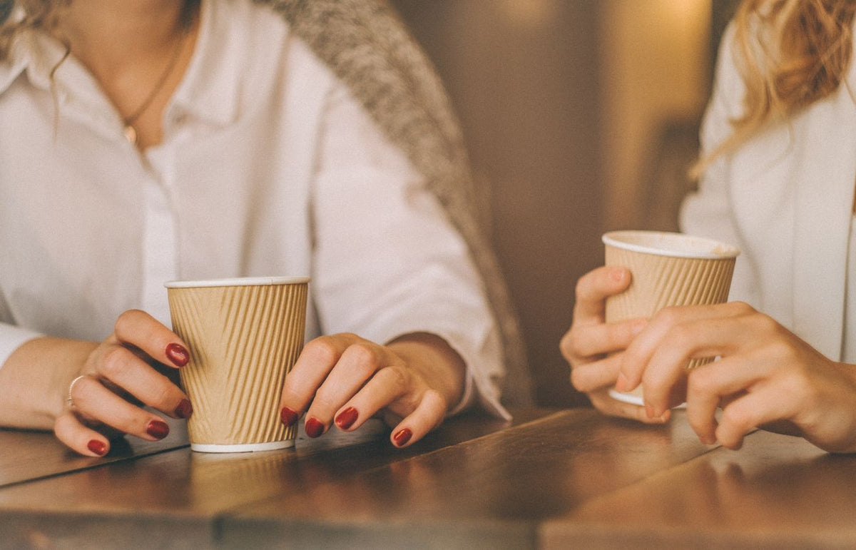 Two people holding paper coffee cups on a wooden table indoors, sharing a moment.