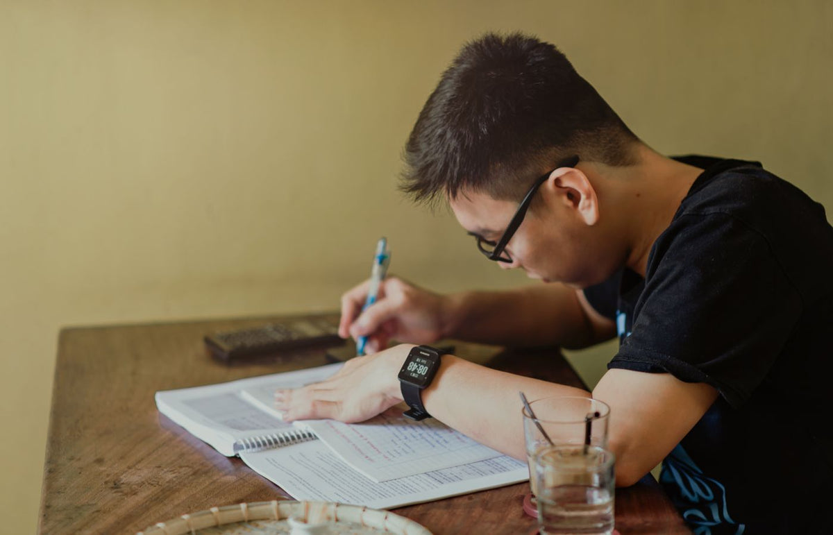 A young man concentrates on studying at his desk, taking notes indoors.