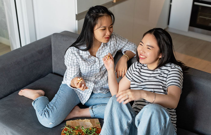 Two friends sit on a sofa sharing pizza, smiling and enjoying a relaxed day indoors.