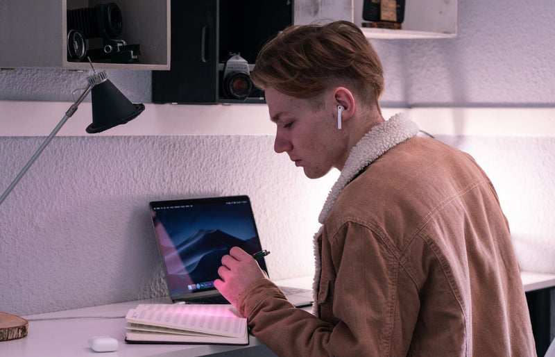 A young man with earbuds focuses on studying using a tablet and notebook in a modern setting.