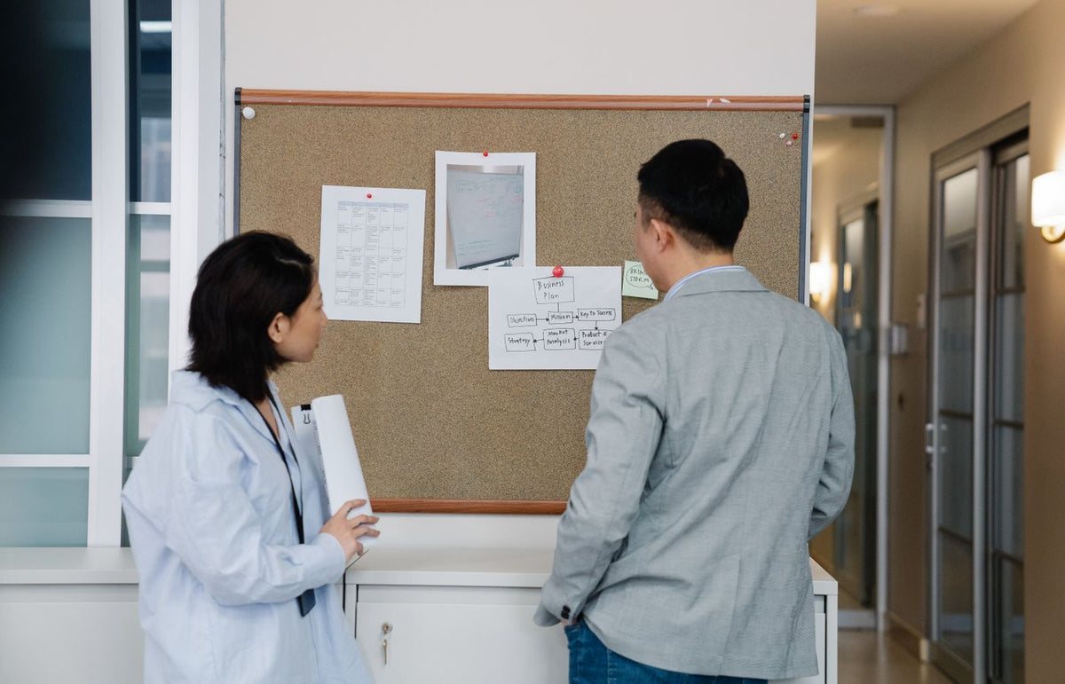 Two coworkers discussing work plans on a noticeboard in a modern office corridor.