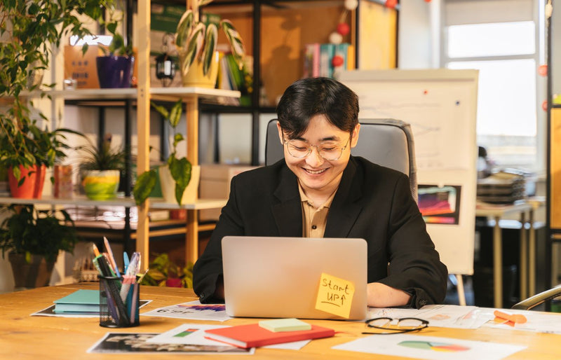 Smiling young professional in black suit working on laptop at a cozy office desk surrounded by plants.