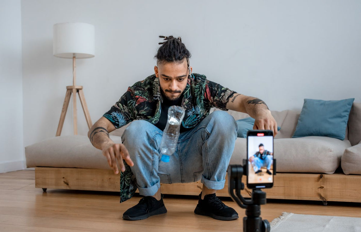 A young man focuses on a bottle flip challenge while filming content at home, capturing it on a smartphone.