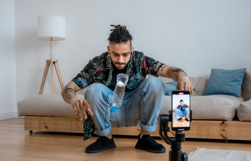 A young man focuses on a bottle flip challenge while filming content at home, capturing it on a smartphone.