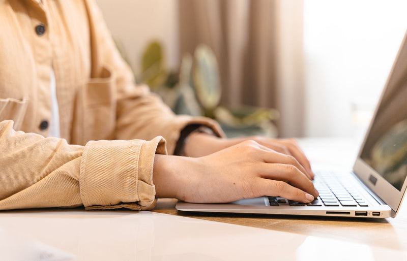 Close-up of hands typing on a laptop indoors, wearing a casual jacket.