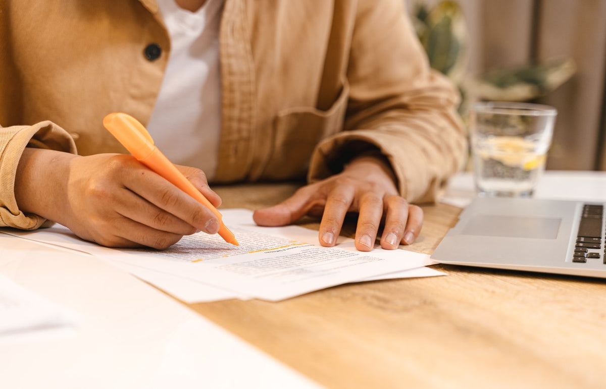 Person highlighting notes at a desk with a laptop and glass of water nearby.