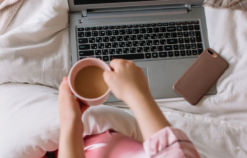 A cozy morning scene with a woman holding coffee, phone, and laptop on bed.